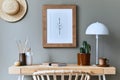 Stylish bohemian interior of home office space with wooden desk, rattan armchair, brown mock up poster frame, cacti.