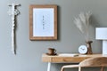 Stylish bohemian interior of home office space with wooden desk, rattan armchair, brown mock up poster frame, cacti.