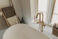 Stylish boho bathroom design. Modern bathtub with golden faucet from floor, wooden ladder with towels and big window, modern eco Royalty Free Stock Photo