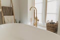 Stylish boho bathroom design. Modern bathtub with golden faucet from floor, wooden ladder with towels, washing essentials and big Royalty Free Stock Photo