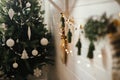Stylish boho christmas tree with modern white baubles on background of garland with bells, rustic stocking and christmas lights.