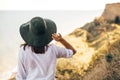 Stylish boho girl in hat looking at sea in sunny evening light from sandy cliff, back view. Happy young hipster woman relaxing on Royalty Free Stock Photo