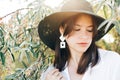 Stylish boho girl in hat and with modern earrings posing among green olive branches in soft evening light. Young fashionable woman Royalty Free Stock Photo
