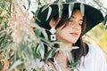 Stylish boho girl in hat and with modern earrings posing among green olive branches in soft evening light. Young fashionable woman