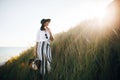 Stylish boho girl in hat posing among grass in sunny evening light at sandy cliff near sea. Happy young fashionable woman relaxing Royalty Free Stock Photo