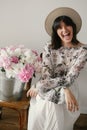 Stylish boho girl in hat sitting and laughing at metal bucket with peonies on rustic wooden chair. Beautiful hipster woman posing Royalty Free Stock Photo