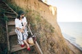 Stylish boho girl in hat sitting on wooden stairs in sunny light at sandy cliff near sea. Happy young fashionable woman relaxing Royalty Free Stock Photo