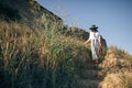 Stylish boho girl in hat walking at sandy cliff with grass near sea, in sunny light. Happy young hipster woman relaxing on Royalty Free Stock Photo