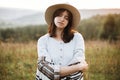 Stylish boho girl portrait in sunny light at atmospheric sunset in meadow. Calm hipster woman in poncho and hat enjoying traveling