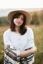 Stylish boho girl portrait in sunny light at atmospheric sunset in meadow. Calm hipster woman in poncho and hat enjoying traveling