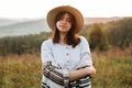 Stylish boho girl portrait in sunny light at atmospheric sunset in meadow. Calm hipster woman in poncho and hat enjoying traveling