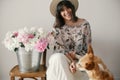 Stylish boho girl sitting at metal bucket with peonies on rustic wooden chair and playing with cute golden dog.Beautiful hipster Royalty Free Stock Photo