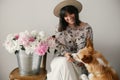 Stylish boho girl sitting at metal bucket with peonies on rustic wooden chair and playing with cute golden dog.Beautiful hipster Royalty Free Stock Photo