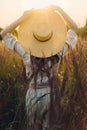 Stylish boho woman with straw hat posing among wild grasses in sunset light, back view. Summer travel. Young carefree female in Royalty Free Stock Photo