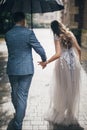 Stylish bride and groom walking under umbrella and holding hands on background of old church in rain. Provence wedding. Beautiful Royalty Free Stock Photo