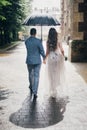 Stylish bride and groom walking under umbrella and holding hands on background of old church in rain. Provence wedding. Beautiful Royalty Free Stock Photo