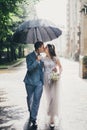 Stylish bride and groom walking under umbrella and kissing on background of old church in rain. Provence wedding. Beautiful Royalty Free Stock Photo