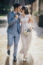 Stylish bride and groom walking under umbrella and kissing on background of old church in rain. Provence wedding. Beautiful Royalty Free Stock Photo