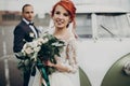 Stylish bride holding modern bouquet and groom looking at her near retro car. luxury wedding couple newlyweds posing. space for Royalty Free Stock Photo