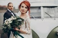 Stylish bride holding modern bouquet and groom looking at her near retro car. luxury wedding couple newlyweds posing. space for Royalty Free Stock Photo