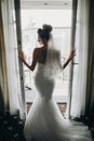 Stylish bride opening window balcony in soft light in hotel room. Back of gorgeous sensual bride in white gown. Morning