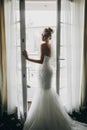 Stylish bride opening window balcony in soft light in hotel room. Back of gorgeous sensual bride in white gown. Morning