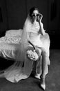 Stylish bride in a wedding dress posing in white glasses sitting on the bed. Black and white photo. Royalty Free Stock Photo