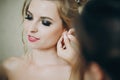 Stylish bridesmaid helping put earring on bride, getting ready in hotel room. Morning preparation before wedding ceremony.