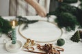 Stylish candle as fir tree, golden bells and ribbon on background of woman hands making boho wreath on white wooden table. Making