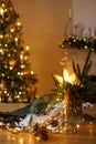 Stylish candle, golden lights, pine cones and ornaments on wooden table against stylish decorated christmas tree and fireplace