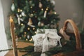 Stylish candle, wrapped christmas gift, rustic basket with fir branches against festive decorated tree in scandinavian room. Moody