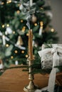 Stylish candle, wrapped christmas gift, rustic basket with fir branches against festive decorated tree in scandinavian room. Moody