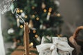 Stylish candle, wrapped christmas gift, rustic basket with fir branches against festive decorated tree in scandinavian room. Moody