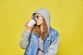 Stylish Caucasian girl in jeans and glasses 8 bit on a yellow background. Holding a disposable coffee cup to go and drinks. Copy Royalty Free Stock Photo