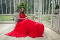 Stylish celebrity woman in red silky dress sitting in white summer pavilion