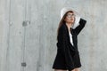 Stylish charming young woman in vintage glasses in fashion black-white clothes and straw hat posing on gray background in street. Royalty Free Stock Photo
