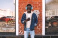 Stylish cheerful african man using smartphone listening to music in wireless headphones on city street over brick wall Royalty Free Stock Photo