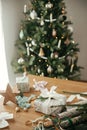 Stylish christmas gift with white ribbon and modern decorations on wooden table on background of festive decorated tree in Royalty Free Stock Photo