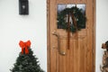 Stylish christmas rustic wreath with vintage bells and ribbon hanging on wooden front door and fir tree with red bow at house