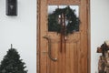 Stylish christmas rustic wreath with vintage bells and ribbon on wooden doors, fir tree and lantern on house exterior. Winter
