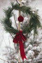 Stylish Christmas wreath and red bauble on tree in snowy winter park. Simple xmas wreath with pine branches and red bow hanging Royalty Free Stock Photo