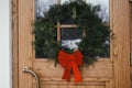 Stylish christmas wreath with red bow hanging on wooden front door at house entrance. Modern minimal farmhouse exterior decor for