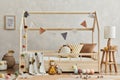 Stylish composition of cozy scandi child`s room interior with wooden bed, pillows, elegant lamp, plush, wooden toys and textile.