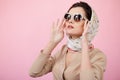 Stylish confident brunette woman touching his sunglasses, wearing in scarves, looking up, isolated on a pink background. Royalty Free Stock Photo