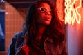 Stylish African woman wears trendy glasses stands near neon sign in night club.