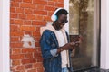 Stylish cool smiling african man using smartphone listening to music in wireless headphones on city street over brick wall Royalty Free Stock Photo