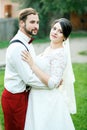 Stylish couple standing embracing, looking to side. Groom with beard, mustache, butterfly tie and suspenders. Bride in