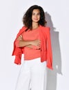 Stylish curly brunette woman in elegant coral shirt, jacket draped over her shoulders and white pants stands