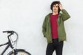 Stylish curly man wears black trousers, green anorak, stands near bicycle against white background, likes riding bike, has active