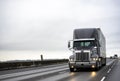 Stylish dark gray big rig semi truck with turned on headlights running with semi trailer on the straight evening wide highway road Royalty Free Stock Photo